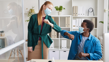 Image of white businesswoman wearing mask bumping elbows with African-American man as a safe greeting in office during COVID-19 pandemic
